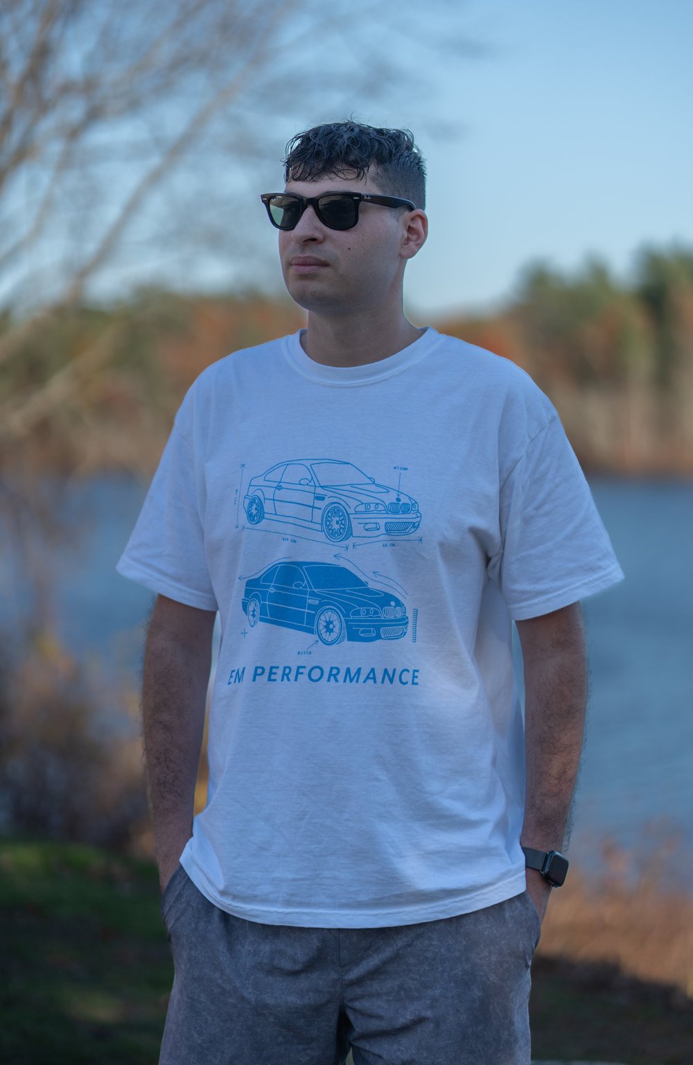 Model wearing E46 M3 graphic t-shirt by E. M. Performance near a lake with trees in background.